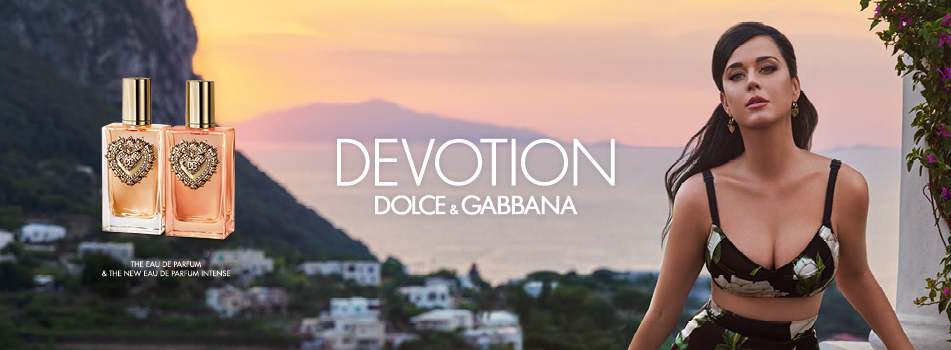 Werbebild für Dolce & Gabbana Devotion: Zwei Parfumflakons mit goldenen Herzverzierungen stehen links vor einer mediterranen Küstenlandschaft bei Sonnenuntergang. Rechts posiert eine Frau in einem geblümten Kleid vor dem malerischen Panorama.
