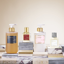 Vier Produkte von Maison Francis Kurkdjian Paris stehen nebeneinander auf einer dekorativen Fläche mit Marmor- und Natursteinoptik. Von links nach rechts: ein oranger Pumpflakon mit dunkelblauem Etikett, ein kleiner Parfümflakon mit rotem Etikett und goldener Kappe, ein weißer Pumpflakon mit rosa Etikett sowie ein kompakter cremefarbener Flakon mit silberner Kappe.