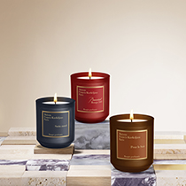 Drei luxuriöse Duftkerzen von Maison Francis Kurkdjian in Rot, Blau und Braun, arrangiert auf dekorativen Marmorblöcken vor hellem Hintergrund.