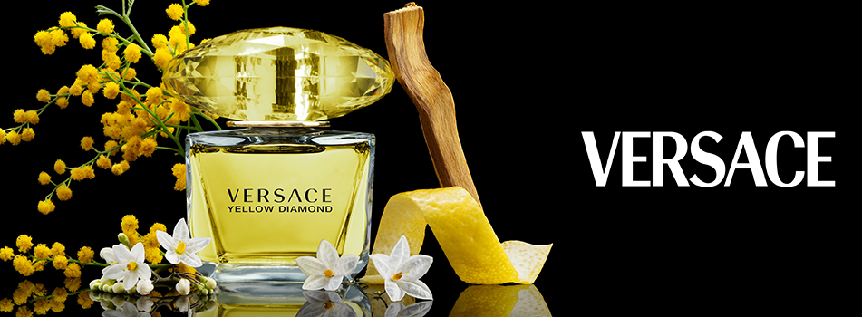 Werbebild des Parfums 'Versace Yellow Diamond'. Der gelbe Flakon mit großem, diamantförmigem Verschluss steht vor schwarzem Hintergrund. Umgeben ist er von gelben Mimosenblüten, weißen Blüten, einem Stück Holz und einer Zitronenschale. Rechts im Bild steht das Versace-Logo in weißer Schrift.